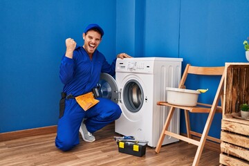 Young hispanic man working on washing machine screaming proud, celebrating victory and success very excited with raised arms