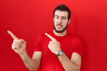 Young hispanic man wearing casual red t shirt pointing aside worried and nervous with both hands,...