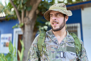 Young hispanic man wearing camouflage army uniform outdoors looking away to side with smile on face, natural expression. laughing confident.