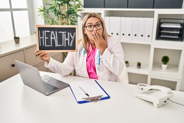 Young hispanic doctor woman holding healthy word covering mouth with hand, shocked and afraid for...