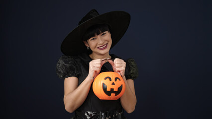 Young chinese woman wearing witch costume holding halloween pumpkin basket over isolated black background