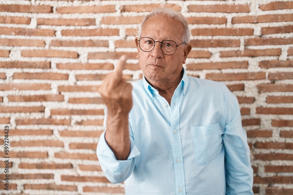 Sticker Senior man with grey hair standing over bricks wall showing middle finger, impolite and rude fuck off expression