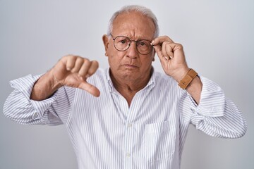 Senior man with grey hair holding glasses with hand with angry face, negative sign showing dislike with thumbs down, rejection concept