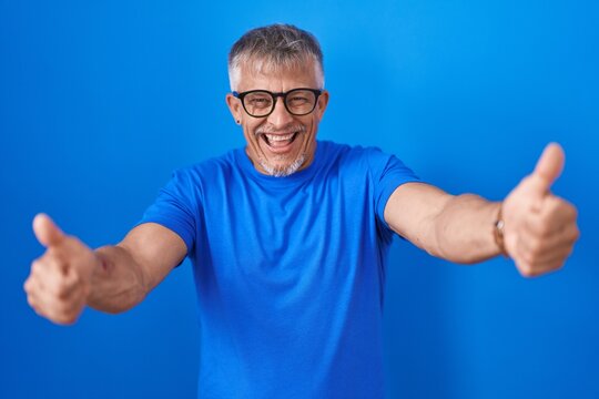 Hispanic man with grey hair standing over blue background approving doing positive gesture with hand, thumbs up smiling and happy for success. winner gesture.