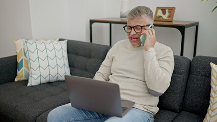 Middle age grey-haired man talking on smartphone using laptop at home