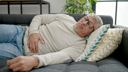 Middle age grey-haired man suffering for stomach ache lying on sofa at home