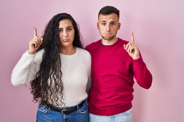 Young hispanic couple standing over pink background pointing up looking sad and upset, indicating...