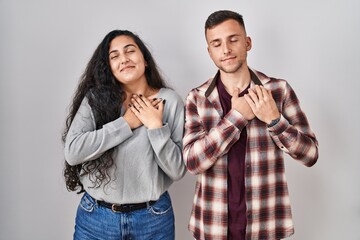 Young hispanic couple standing over white background smiling with hands on chest with closed eyes and grateful gesture on face. health concept.