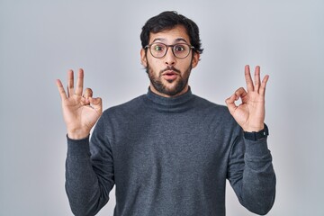 Handsome latin man standing over isolated background looking surprised and shocked doing ok approval symbol with fingers. crazy expression