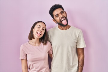 Fototapeta premium Young hispanic couple together over pink background sticking tongue out happy with funny expression. emotion concept.