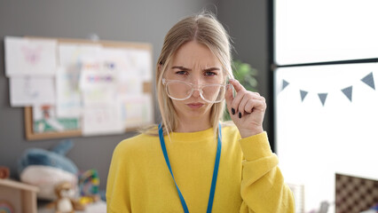 Young blonde woman preschool teacher with angry face holding glasses at kindergarten