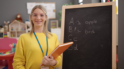 Young blonde woman preschool teacher writing vocabulary exercise on blackboard at kindergarten