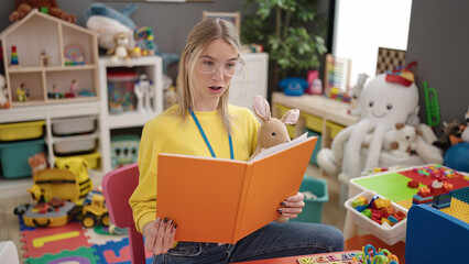 Young blonde woman preschool teacher reading book out loud at kindergarten