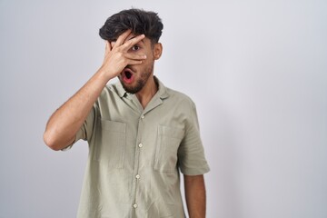 Arab man with beard standing over white background peeking in shock covering face and eyes with...