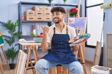 Arab man with beard painter sitting at art studio holding palette pointing thumb up to the side smiling happy with open mouth