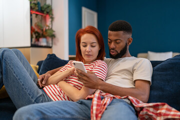 Young millennial diverse couple man and woman resting at home with mobile phone, hugging and surfing internet while relaxing together on sofa in living room. Family bonding and smartphone