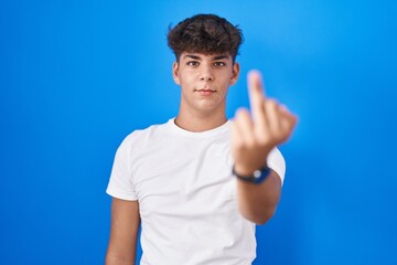 Hispanic teenager standing over blue background showing middle finger, impolite and rude fuck off expression - obrazy, fototapety, plakaty