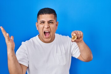 Fototapeta premium Hispanic young man holding virtual currency bitcoin celebrating victory with happy smile and winner expression with raised hands