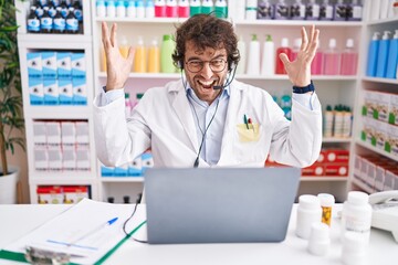 Hispanic young man working at pharmacy drugstore working with laptop celebrating victory with happy smile and winner expression with raised hands