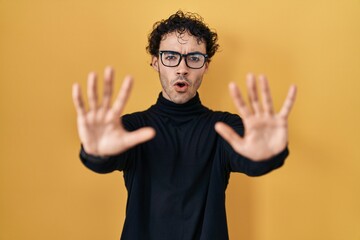 Hispanic man standing over yellow background doing stop gesture with hands palms, angry and frustration expression