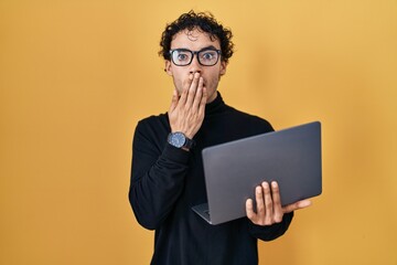 Hispanic man working using computer laptop covering mouth with hand, shocked and afraid for mistake. surprised expression