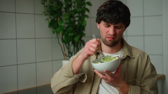 A man in a beige shirt eats a tasteless salad in the kitchen. 