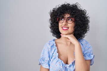 Young brunette woman with curly hair wearing glasses over isolated background with hand on chin thinking about question, pensive expression. smiling with thoughtful face. doubt concept.
