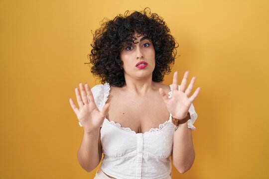 Young brunette woman with curly hair standing over yellow background moving away hands palms showing refusal and denial with afraid and disgusting expression. stop and forbidden.
