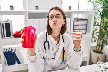 Young caucasian dentist woman holding denture and mouthwash looking at the camera blowing a kiss being lovely and sexy. love expression.