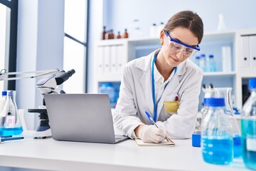 Young caucasian woman scientist using laptop writing on notebook at laboratory