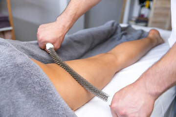 A chiropractor uses a metal tool to massage the leg of a patient who is lying on a comfortable couch