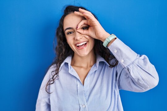 Young brunette woman standing over blue background doing ok gesture with hand smiling, eye looking through fingers with happy face.