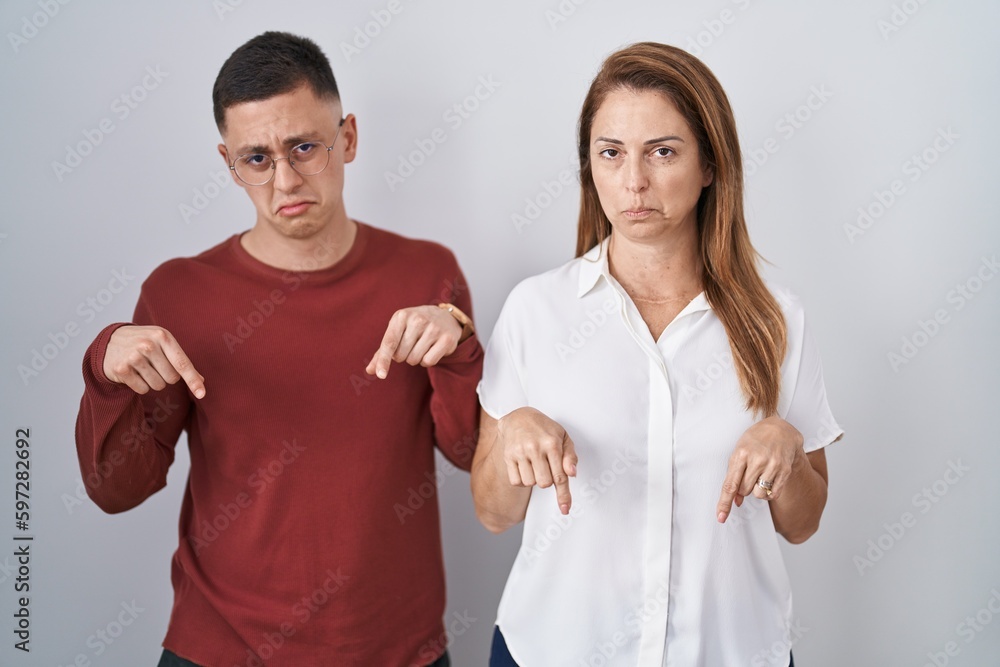 Sticker Mother and son standing together over isolated background pointing down looking sad and upset, indicating direction with fingers, unhappy and depressed.