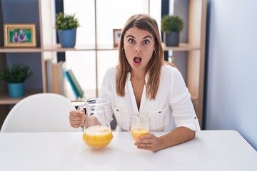 Young hispanic woman drinking glass of orange juice afraid and shocked with surprise and amazed expression, fear and excited face.