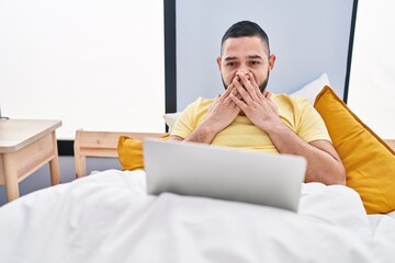 Hispanic man using laptop on the bed covering mouth with hand, shocked and afraid for mistake....