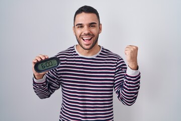 Young hispanic man wearing pyjama holding alarm clock screaming proud, celebrating victory and success very excited with raised arms