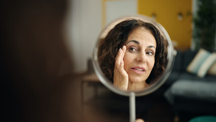 Middle age hispanic woman sitting on sofa looking face on mirror at home