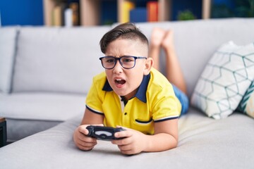 Young hispanic kid playing video game holding controller on the sofa angry and mad screaming...