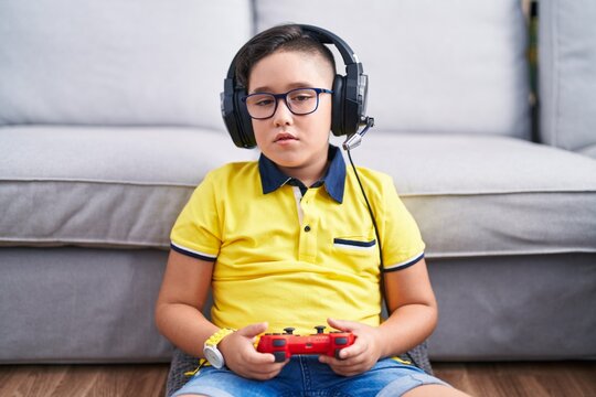 Young hispanic kid playing video game holding controller wearing headphones looking sleepy and tired, exhausted for fatigue and hangover, lazy eyes in the morning.