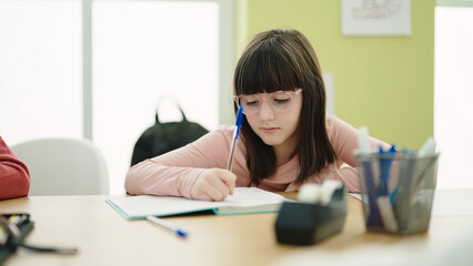 Adorable hispanic girl student writing on notebook at classroom