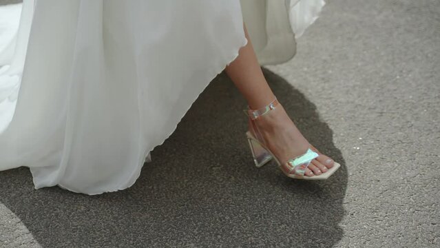 Slender legs of bride in translucent iridescent mother of pearl shoes walk along paved road with markings. Walking in white dress on sunny hot day, close up.