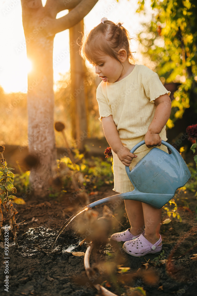 Wall mural cute little baby girl watering flowers garden on sunny day from a watering can. family home leisure.