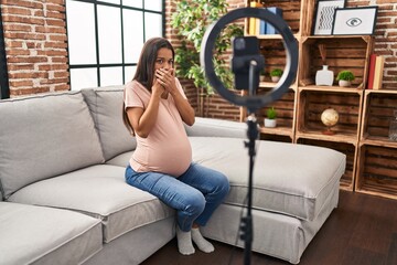 Fototapeta na wymiar Young pregnant woman recording vlog tutorial with smartphone at home shocked covering mouth with hands for mistake. secret concept.