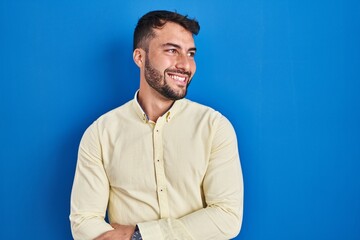 Obraz premium Handsome hispanic man standing over blue background looking away to side with smile on face, natural expression. laughing confident.