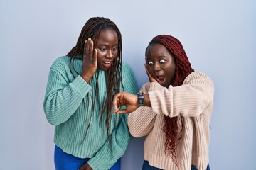 Two african woman standing over blue background looking at the watch time worried, afraid of...