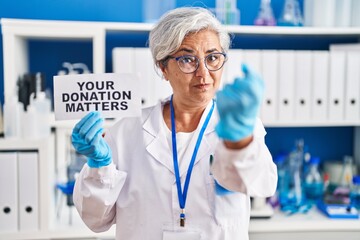 Middle age woman with grey hair working at scientist laboratory holding your donation matters banner annoyed and frustrated shouting with anger, yelling crazy with anger and hand raised