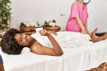 Obraz na płótnie Canvas Young african american woman holding credit card having legs massage at beauty center