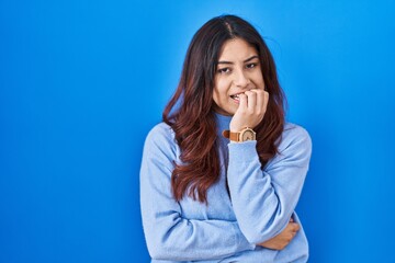 Hispanic young woman standing over blue background looking stressed and nervous with hands on mouth biting nails. anxiety problem.