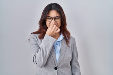 Hispanic young business woman wearing glasses smelling something stinky and disgusting, intolerable smell, holding breath with fingers on nose. bad smell