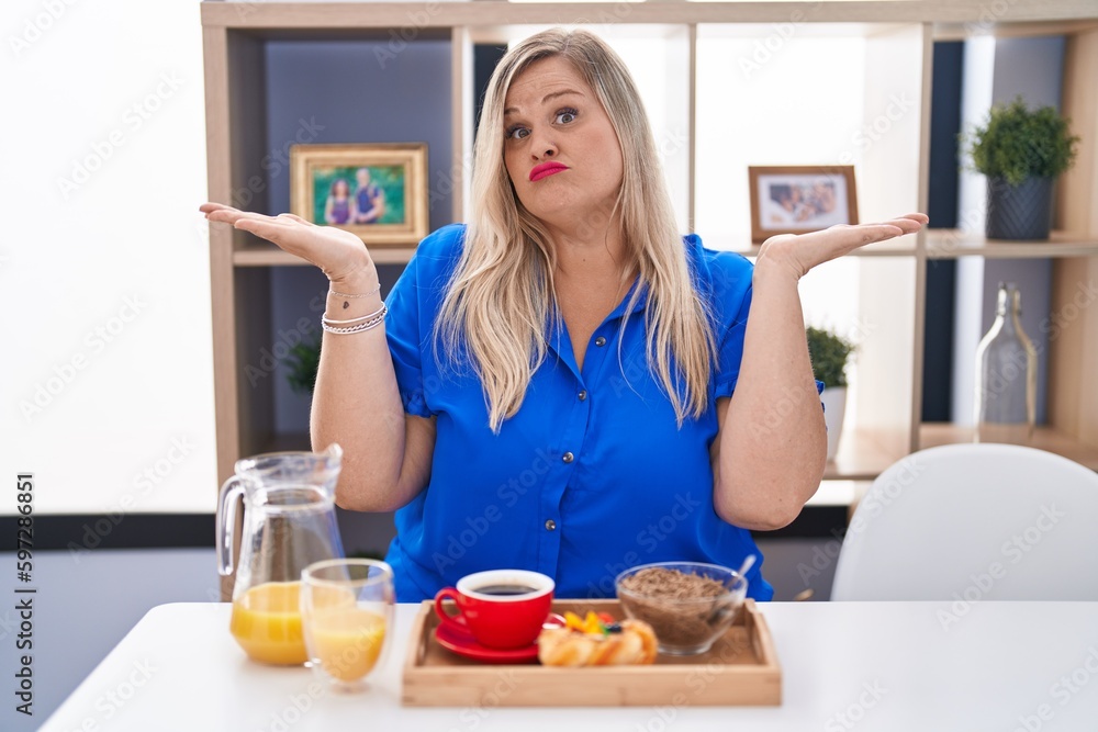 Sticker caucasian plus size woman eating breakfast at home clueless and confused expression with arms and ha